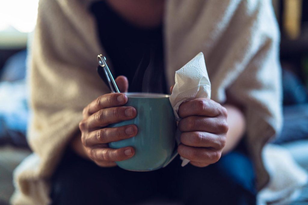 Individual with the flu, wrapped in a blanket and drinking a warm cup tea.